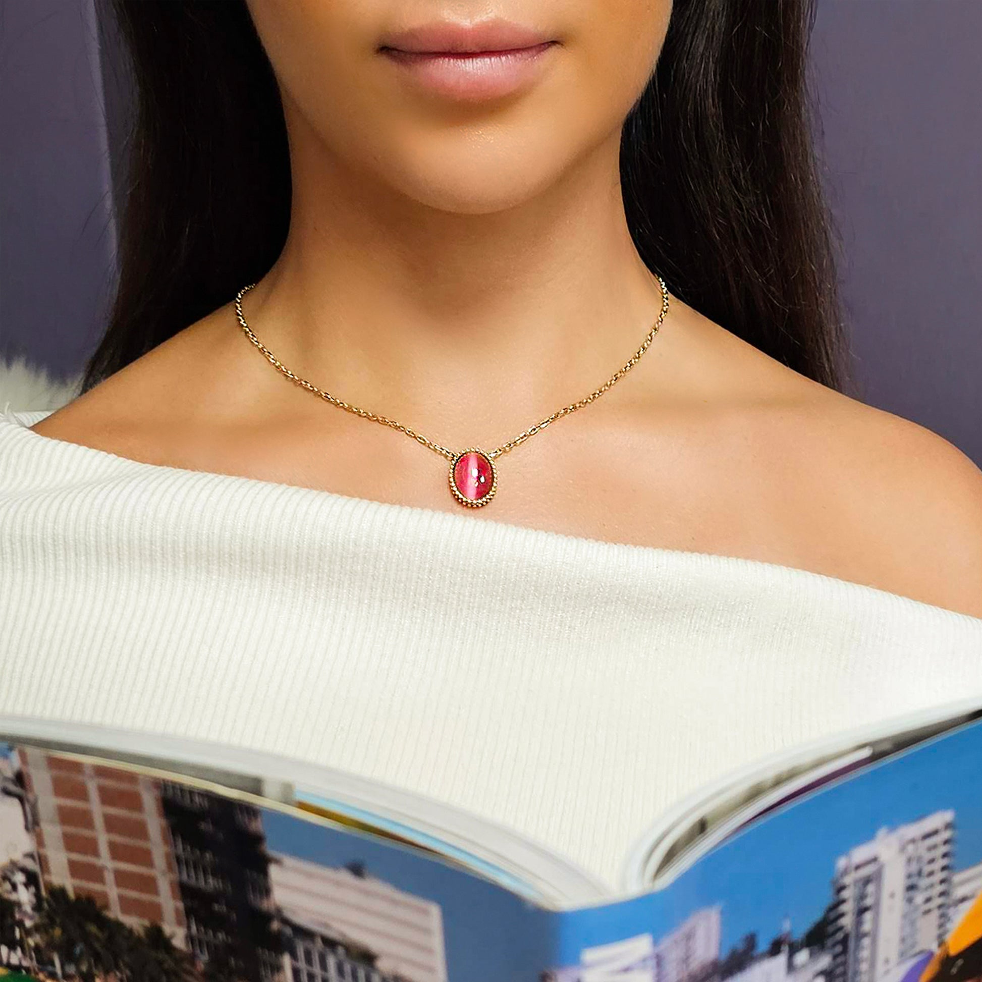 Close-up of a woman wearing a gold necklace with a red pendant, against a blurred background.