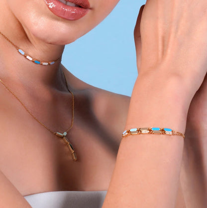 Close-up of a woman wearing a gold necklace and bracelet with blue stones against a light blue background.