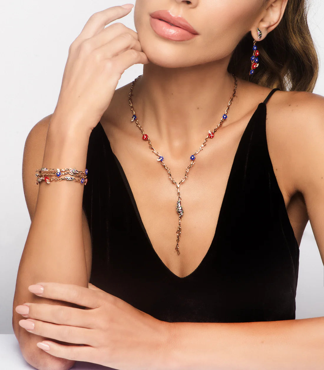 Woman wearing a black top with jewelry including a necklace, bracelet, and earrings on a light gray background.