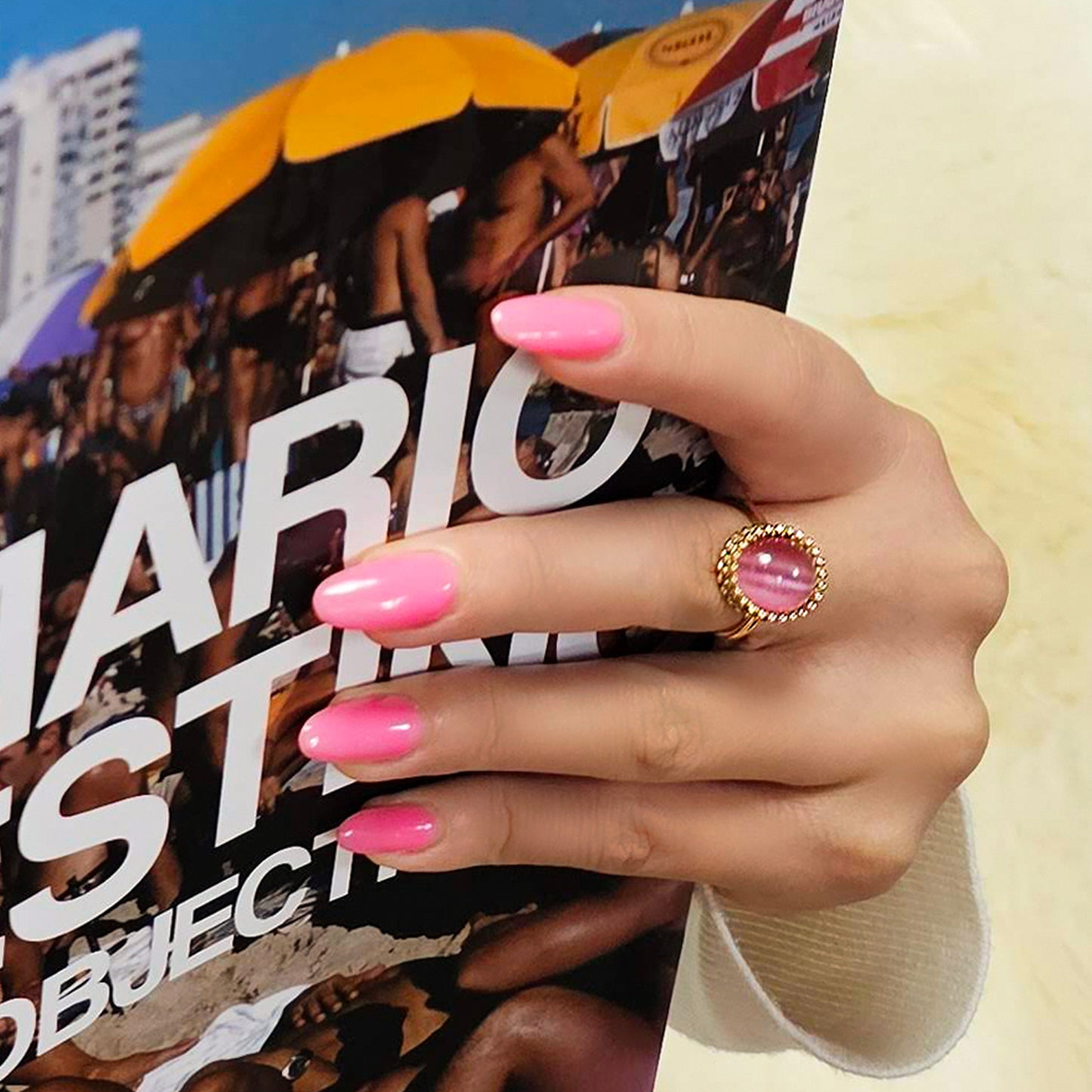 Hand with pink nails holding a magazine featuring a fashion model against a cityscape. Gold ring with a pink gemstone_2
