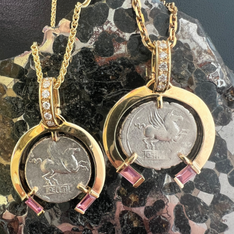 Two gold necklaces with circular pendants featuring coins and pink gemstones on a dark stone surface.