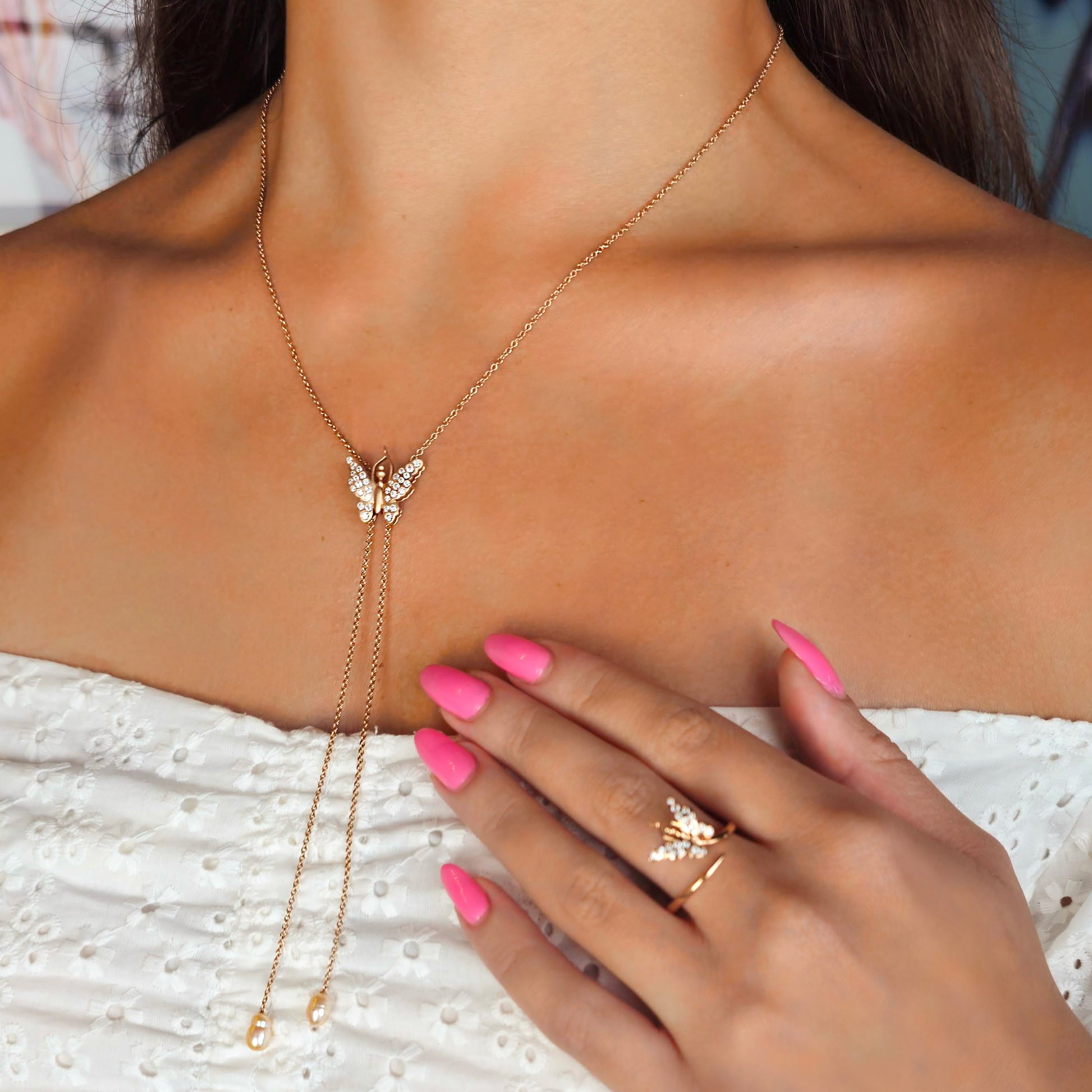 Gold butterfly necklace on a person's neck with a hand wearing a ring, against a blurred background.