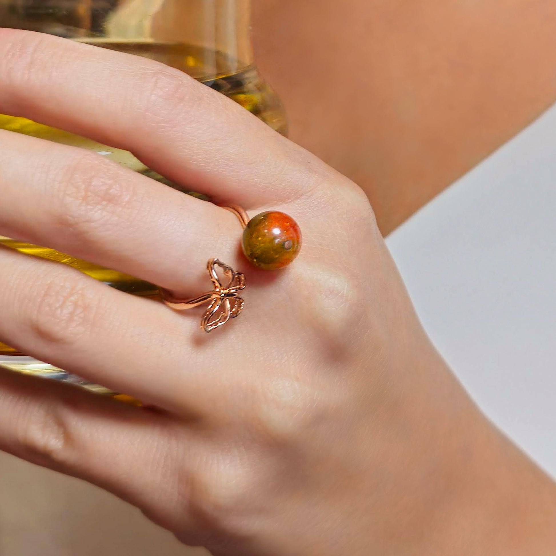 Hand wearing a ring with a red and orange stone, holding a glass of yellow liquid.