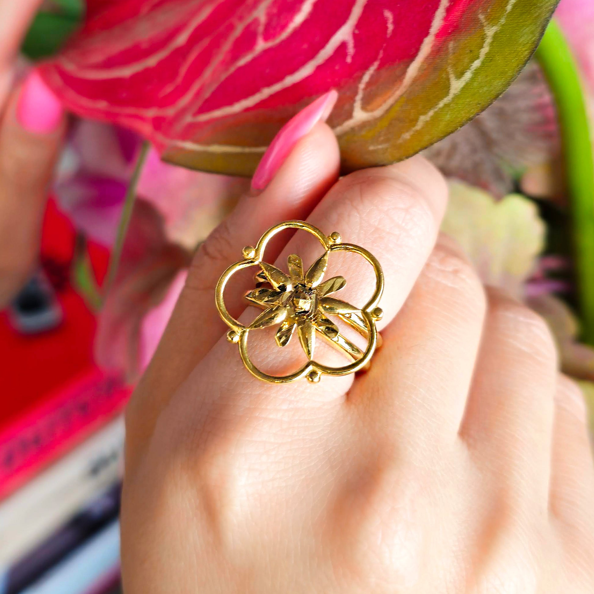 Gold ring with a floral design worn on a finger, held against a blurred background of flowers.