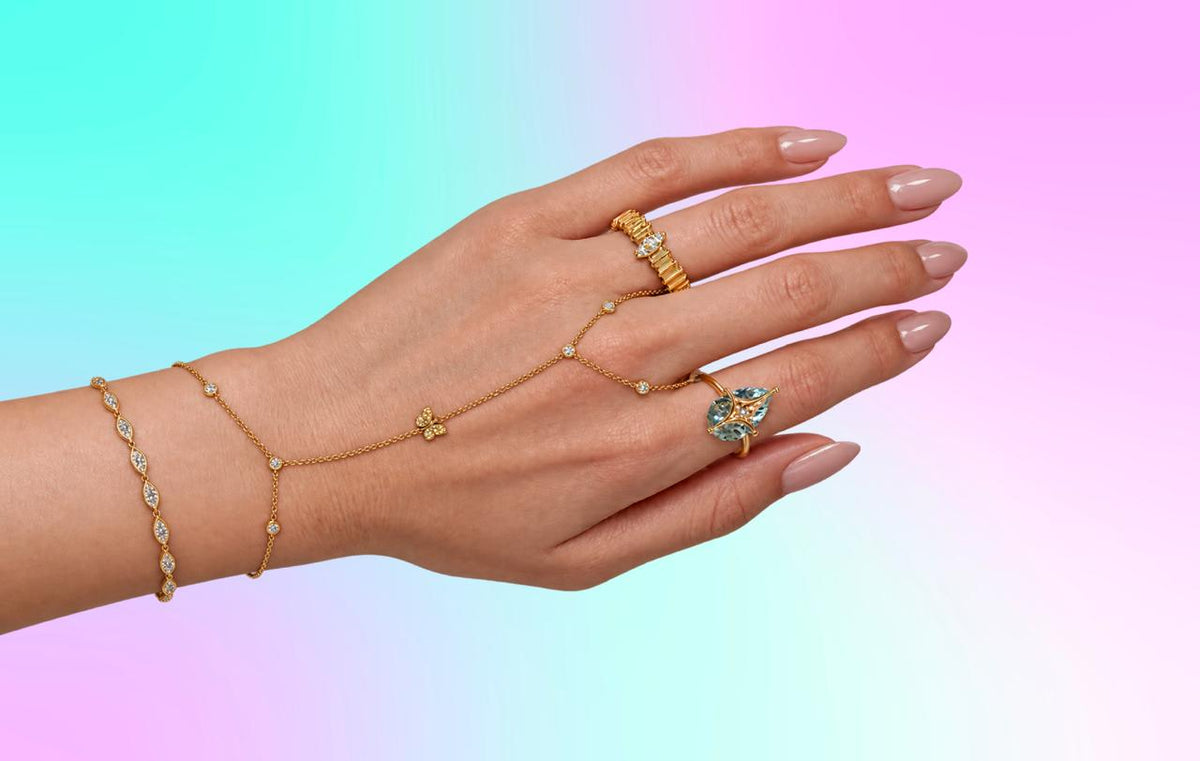 Full hand wearing JOOLS gold bracelets, hand chain and gemstone rings against a pastel background