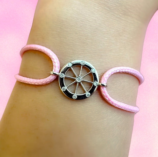Pink bracelet with a gold wheel charm on a wrist against a pink background